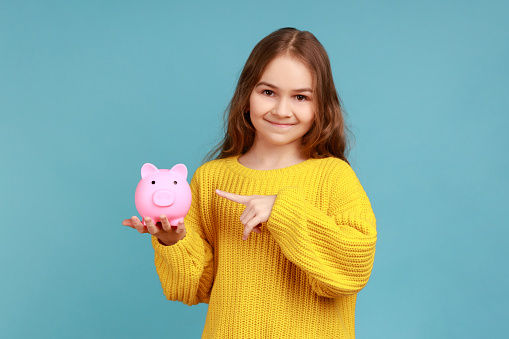 Little girl holding and pointing at piggy bank with finger, suggesting you to put coins inside, wearing yellow casual style sweater. Indoor studio shot isolated on blue background.