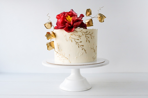 White wedding cake decorated with red flower and golden leaves on a white background.