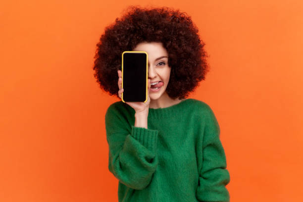 Positive funny woman with Afro hairstyle wearing green casual style sweater covering eye with cell phone with blank screen and showing tongue out. stock photo