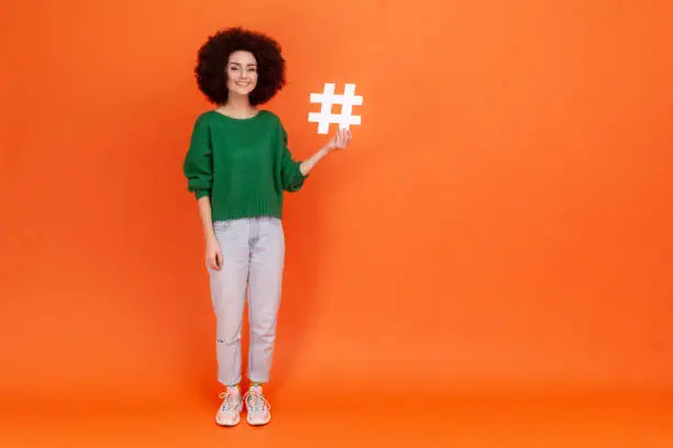 Photo of Full length portrait of woman with Afro hairstyle wearing green casual style sweater presenting white hashtag symbol, internet popularity.