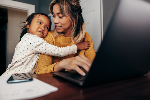 Adorable little girl embracing her mom in her home office. Working mom smiling at her daughter while typing an email on a laptop. Self-employed mother of one working remotely.