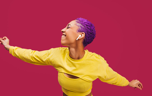 Vibrant dancing. Happy young woman dancing with her arms outstretched while wearing wireless earphones in a studio. Woman with purple hair streaming her favourite music playlists.