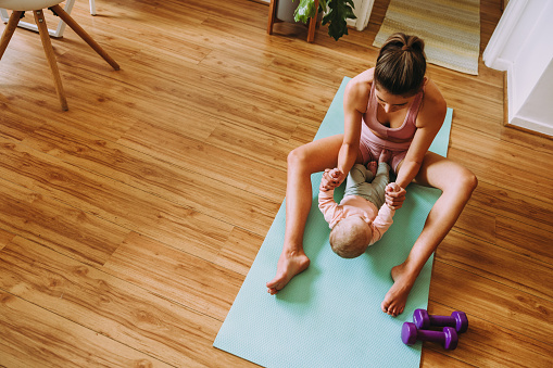 Overhead view of a mom working out with her baby on an exercise mat. Caring mom doing sit up exercises with her baby at home. New mom bonding with her baby during her post-natal fitness routine.
