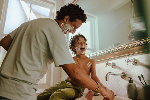 Cheerful father and son having fun with shaving foam in the bathroom. Father and son laughing happily with shaving cream on their faces. Loving father bonding with his young son at home.