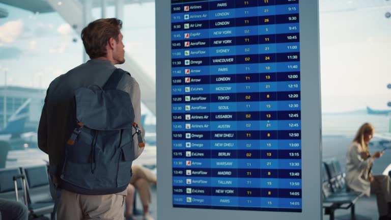 Airport Terminal: Young Man Looking at Arrival and Departure Information Display Looking for His Flight. Backgrond: Diverse Crowd of People Wait for their Flights in Boarding Lounge of Airline Hub