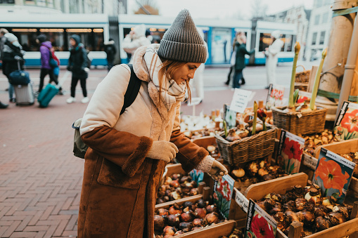 Young woman wearing a hat, coat, and a backpack buying tulip bulbs in a street vendor in Amsterdam city during her vacation in the Netherlands