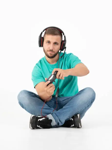 Young man playing videogames with a controller and a headphones in a white background with jeans, black shoes and green shirt