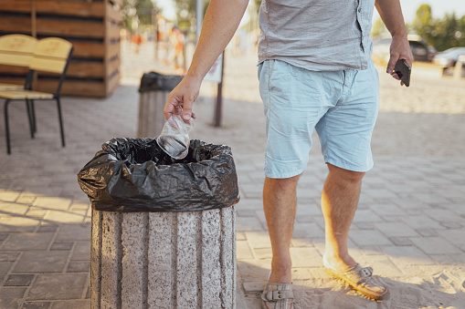 man in summer shorts and T-shirt throws plastic cup into trash can keeping clean outside beach cafe