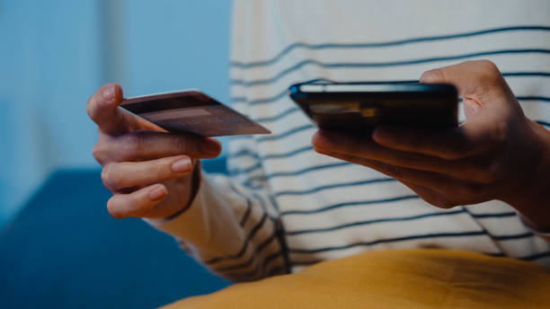 istock Young Asian woman using phone while performing online transactions with credit card during shopping sitting on sofa in living room at home at night. 1362907626