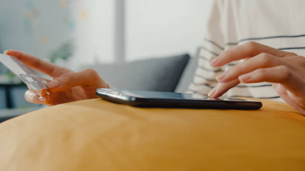 istock Young Asian woman using phone while performing online transactions with credit card during shopping sitting on sofa in living room at home. 1362909239