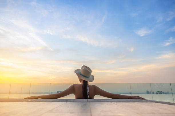 back view of young woman in bikini with straw hat on the sun-tanned slim, shapely body with her arms spread to the side, relaxing in swimming pool on the roof top of hotel, enjoy cityscape at sunset. - zwembad fotos stockfoto's en -beelden