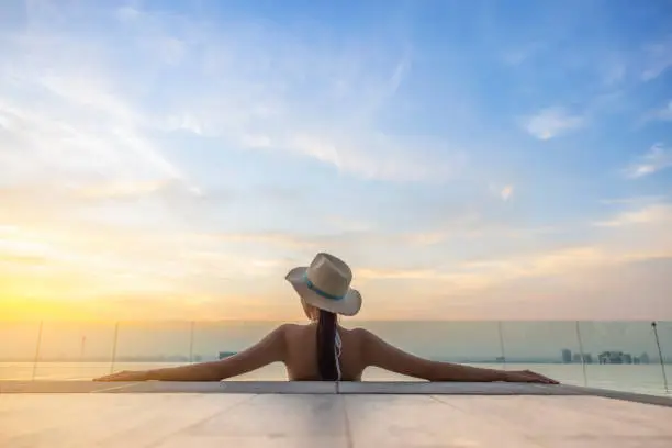 Photo of Back view of young woman in bikini with straw hat on the sun-tanned slim, shapely body with her arms spread to the side, relaxing in swimming pool on the roof top of hotel, enjoy cityscape at sunset.