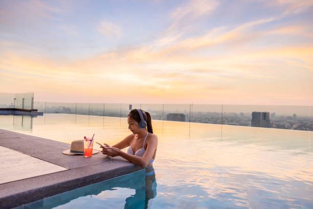 estilo de vida de moda de la mujer asiática en bikini disfrutando de buena música en auriculares a través de la aplicación en el teléfono inteligente cerca de la piscina en sus vacaciones. cóctel de fresa con sombrero en el borde de la piscina. ciuda - swimwear bikini swimming pool red fotografías e imágenes de stock