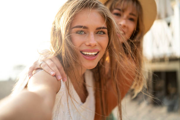 dos felices hermanas caucásicas rubias divirtiéndose durante el verano, tomándose selfies, sonriendo y mirando la cámara del teléfono inteligente. - on the phone women beautiful beauty fotografías e imágenes de stock