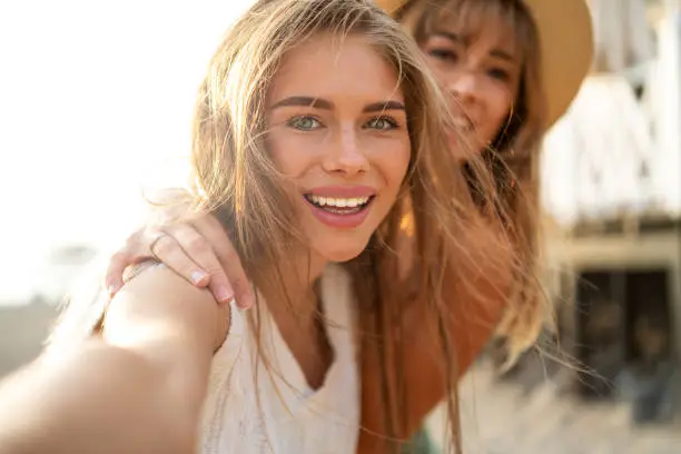 Photo of Two happy blonde caucasian sisters having fun during summer, taking selfie, smiling and looking to the smartphone camera.