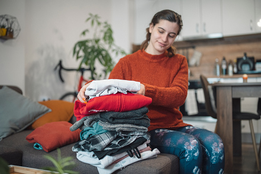 Stack of clean freshly laundered, neatly folded clothes on wooden table. Pile of shirts, dresses and sweaters on the table, white wall background. Copy space.