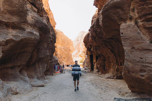 Young man explorer walking inside the canyon looking at the old ruins of Petra town in the Middle East