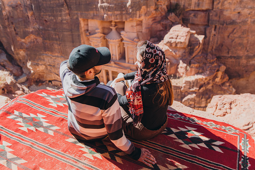 Young multiracial couple sitting on the top of the cliff looking at the famous old Petra town gates hidden in the canyon in Jordan, the Middle East
