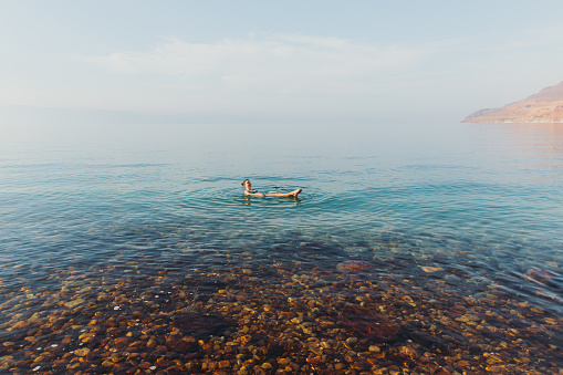 Young woman contemplating the sunny day in the Middle East, swimming in the salty standing waters of the beautiful reflecting sea with mountain view