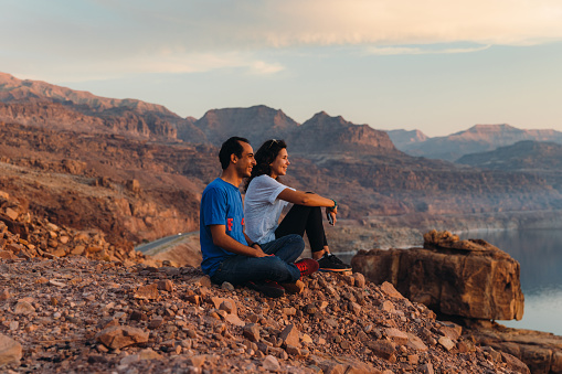 Smiling multiracial couple of travelers feeling freedom and happiness sitting on the mountain peak with dramatic view of the reflection sea from above during bright yellow sunset in the Middle East