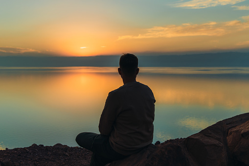 Silhouette of a man traveler feeling freedom and happiness sitting on the mountain peak with dramatic view of the reflecting sea from above during bright yellow sunset in the Middle East