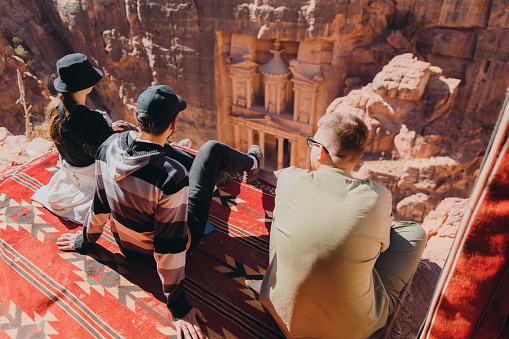 Happy young two multiracial males and one female explorers sitting on the top of the cliff looking at the famous old Petra town gates hidden in the canyon in Jordan, the Middle East