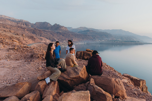 Young multiracial group of females and males feeling freedom and happiness sitting on the mountain peak with dramatic view of the reflection sea from above during bright yellow sunset in the Middle East
