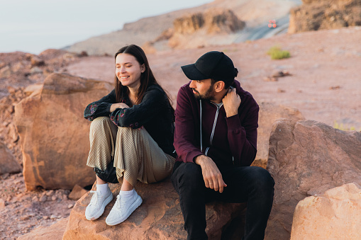 Smiling multiracial couple of travelers feeling freedom and happiness sitting on the mountain peak with dramatic view of the sea from above during bright yellow sunset in the Middle East