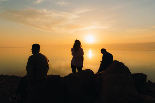 amigos viajantes contemplando o pôr do sol cênico acima do mar morto na jordânia - silhouette three people beach horizon - fotografias e filmes do acervo