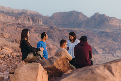 Young multiracial group of females and males feeling freedom and happiness sitting on the mountain peak with dramatic view of the reflection sea from above during bright yellow sunset in the Middle East