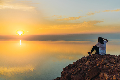Female traveler feeling freedom and happiness sitting on the mountain peak with dramatic view of the reflection sea from above during bright yellow sunset in the Middle East
