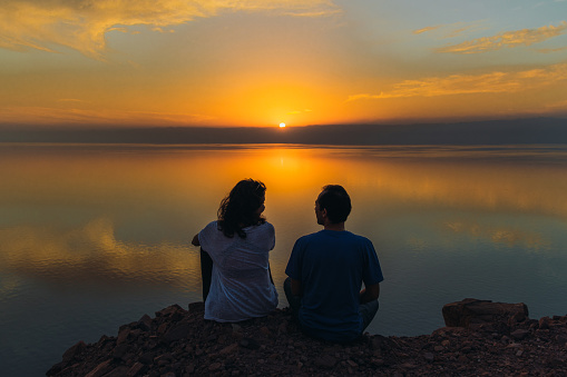 Young multiracial couple of travelers feeling freedom and happiness sitting on the mountain peak with dramatic view of the reflection sea from above during bright yellow sunset in the Middle East