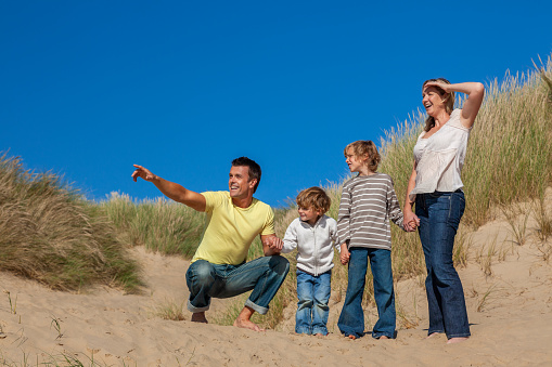 Happy family of mother, father and two sons, man woman children walking holding hands, pointing and having fun in the sand dunes of a sunny beach