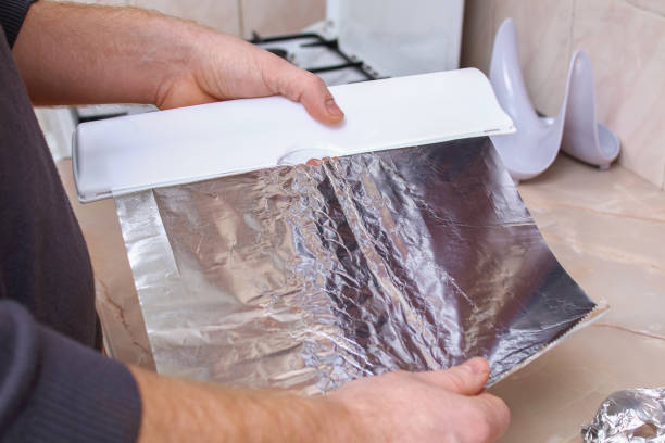 un hombre arranca el papel de aluminio del dispensador de plástico para envolver las papas en él y hornearlas en el horno. - rolled up foil paper gray fotografías e imágenes de stock