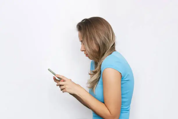 A young blonde woman in a blue t-shirt looks at the mobile phone screen isolated on a white background. Incorrect back position