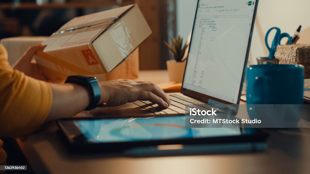Young freelance Asian woman using laptop packing cardboard box in living room at home at night. Working from home concept. Young freelance Asian woman using laptop packing cardboard box in living room at home at night. Working from home. E-commerce Stock Photo