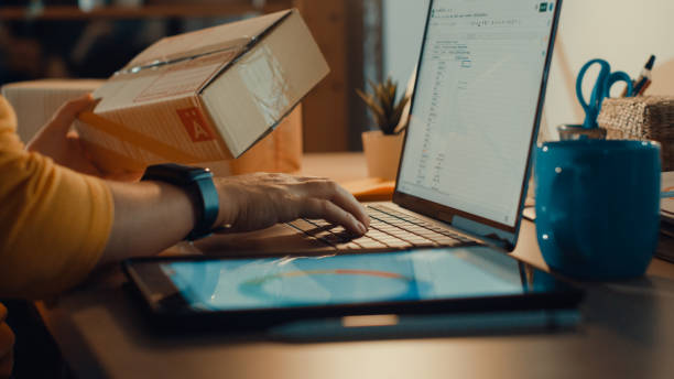 Young freelance Asian woman using laptop packing cardboard box in living room at home at night. Working from home concept. Young freelance Asian woman using laptop packing cardboard box in living room at home at night. Working from home. electronic commerce stock pictures, royalty-free photos & images