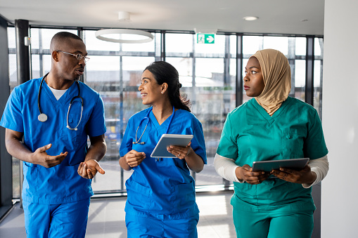 Mixed ethnic group of medical professionals walking down a corridor together in the North East of England. They are working a shift at a hospital and are dressed in scrubs. The women are carrying/using digital tablets.