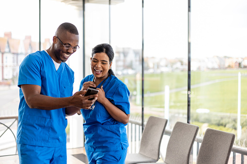 Mixed ethnic group of medical professionals laughing together while looking at a phone in the North East of England. They are working a shift at a hospital and are dressed in scrubs.