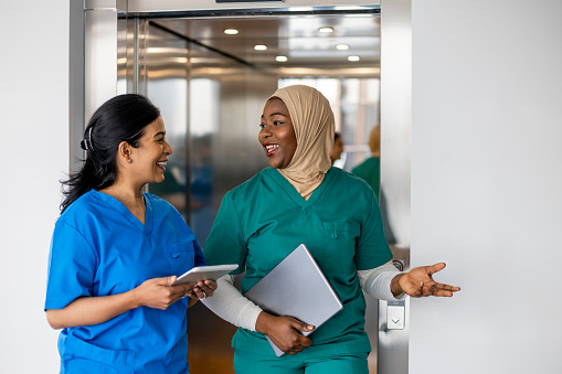 Mixed ethnic group of women medical professionals walking down a corridor together in the North East of England. They are working a shift at a hospital and are dressed in scrubs. They are carrying digital tablets.