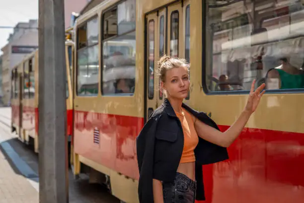 Beautiful natural blonde in a short orange T-shirt, blue jacket and black shorts walking around the city and posing for a photographer as he stops - waving his hand at the city trolleybus
