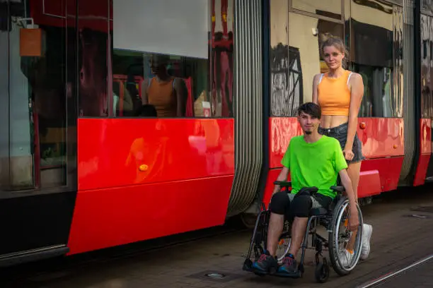 Slender, long-legged natural blonde and a short orange sleeveless T-shirt and black shorts, with a disabled boy in a wheelchair, walking around the city. Portrait next to a passing city bus, trolleybus.