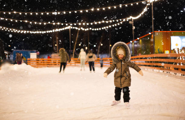 Little girl is skating on ice skates on skating rink in evening, decorated with fairy lights, Christmas trees and fir. Festive mood, Christmas, New Year, holidays, active winter sports and lifestyle stock photo