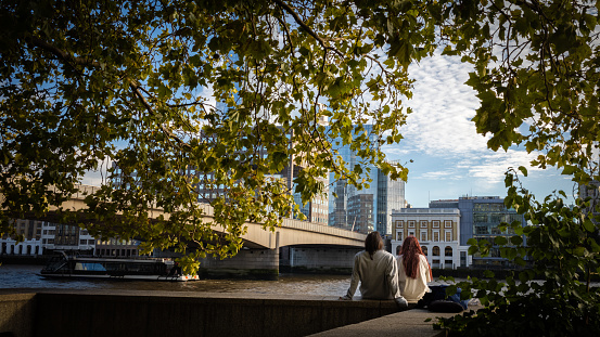 LONDON, UK - October 6, 2021. A young couple sits on the River Thames embankment next to London Bridge in the city centre as a tourist boat passes.