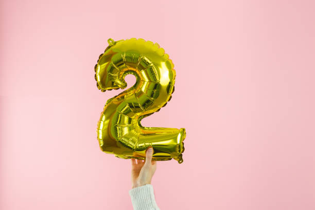 женская рука в белом джемпере держит золотой глобус в форме цифры 2 на розовом фоне. копирование пространства. - puffed sleeve стоковые фото и изображения