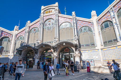 People walking around the entrance to Mercado Central. The exterior of Mercado Central in Valencia. This is a free to enter public modernist building, built between 1914–1928, one of the largest fresh food markets in Europe. The style is variously described as eclecticism and Valencian art nouveau.