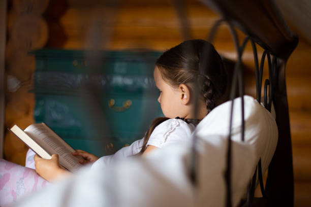 istock Young reading girl side captured from behind bed lying on pillow holding book in hands looking at book reading with dresser in background in wooden house 1362959855