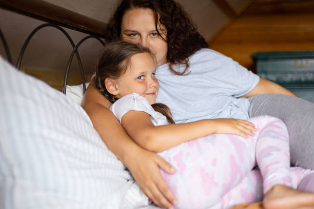 istock Young Caucasian daughter and mother hugging each other lying on double bed both looking at camera in wooden house wearing pajamas in daytime. Family time 1362960481
