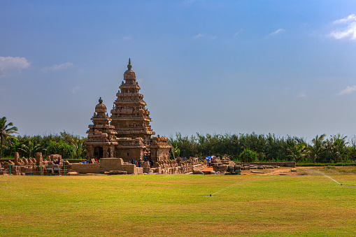 Mahabalipuram, India - January 30, 2015: Tourists at the Shore Temple. located on the shores of the Bay of Bengal, in Mahabalipuram or Mamallapuram, built in the 8th Century, between 700 and 728 AD, during the rign of the Pallava King Narasimhavarman II. Declared a UNESCO World Heritage site in 1984, the temple is one of the oldest structural temples in South India. It survived the Tsunamis of the 13th Century and 2004. Photo shot around noon against a blue sky with some clouds. Copy space.