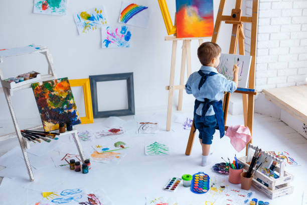 かわいい、深刻で集中した、青いシャツとジーンズのエプロンを着た白い3歳の男の子が、イーゼルの上に立ってキャンバスに描いています。幼児教育、才能、幸せな家族や子育ての概念 - artists canvas indoors childhood small ストックフォトと画像